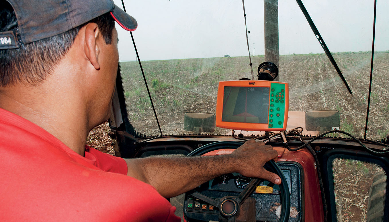 Fotografia. Destaque para um homem de costas, dirigindo um trator, vestindo boné e camiseta vermelha. O homem segura o volante e observa a tela de um GPS. À frente do trator, uma superfície ocupada por plantação.