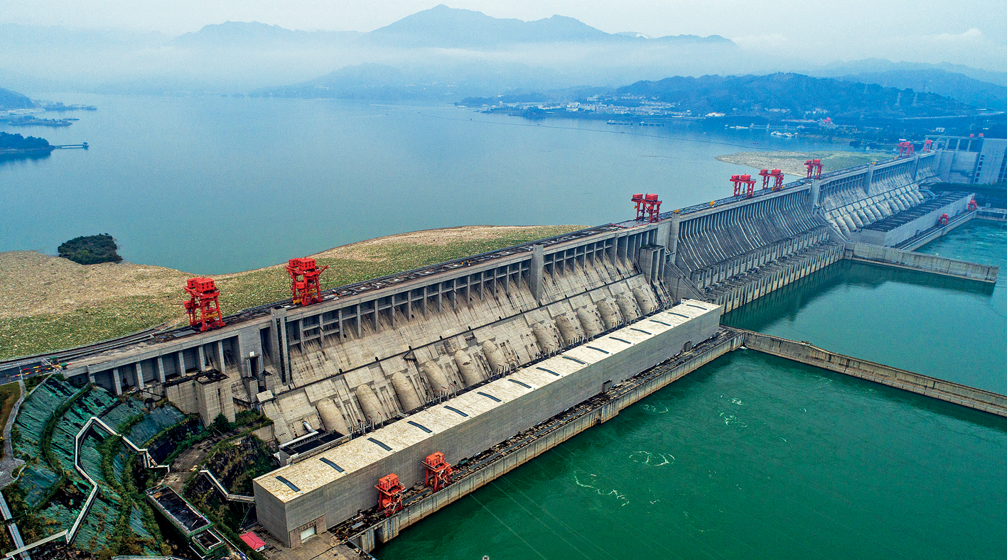 Fotografia. Vista de uma hidrelétrica. No primeiro plano, grande barragem de concreto atravessando uma grande porção de água, dividindo-a. Ao lado direito, a jusante do rio. No segundo plano, o reservatório da hidrelétrica e as margens ocupadas por morros e montanhas.