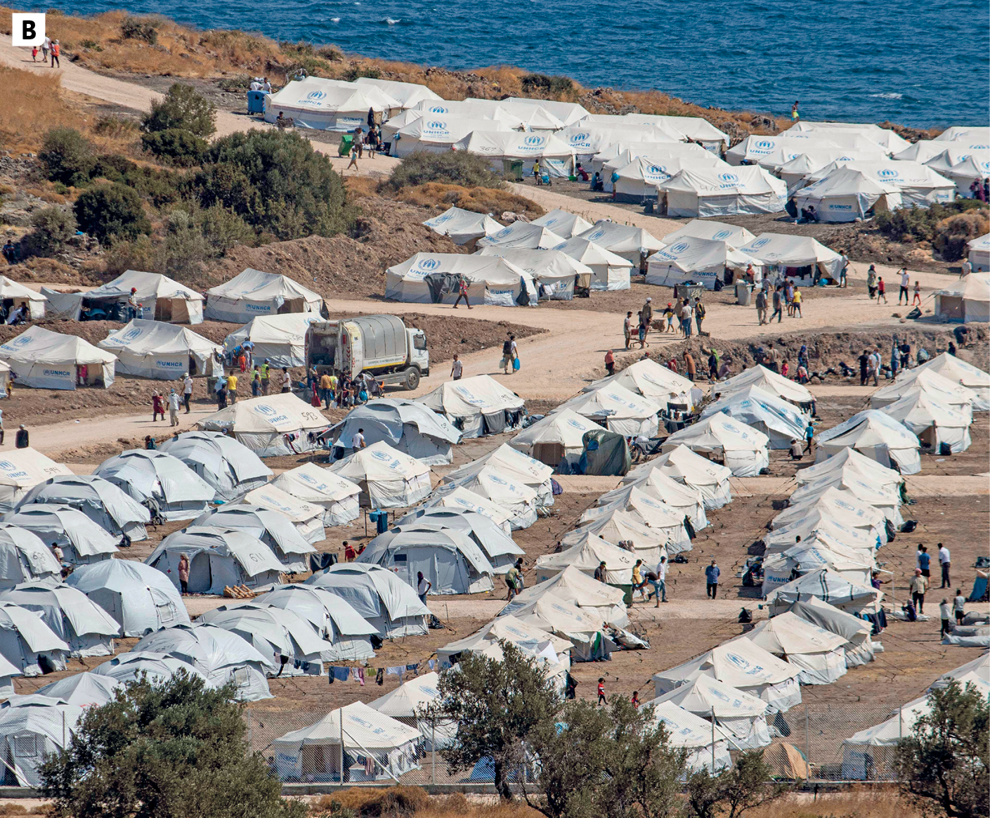 Fotografia B. Vista para um campo de refugiados situado à beira-mar. No primeiro plano, diversas tendas brancas dispostas lado a lado e pessoas circulado entre elas. Algumas árvores esparsas e via de circulação de veículos sem asfalto. No segundo plano, o mar.