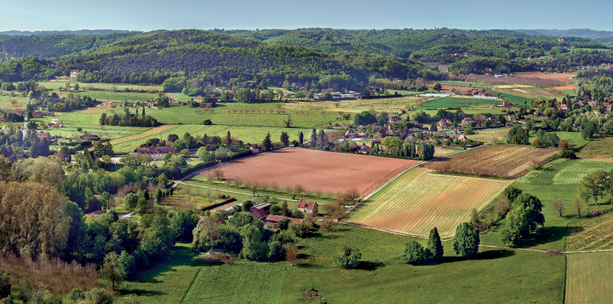 Fotografia. Vista aérea para uma área rural composta por diversos lotes com tamanhos e tipos de cultivos distintos. Entre os lotes, há algumas casas e árvores esparsas. Ao fundo, colinas cobertas por floresta. Acima, céu azul.