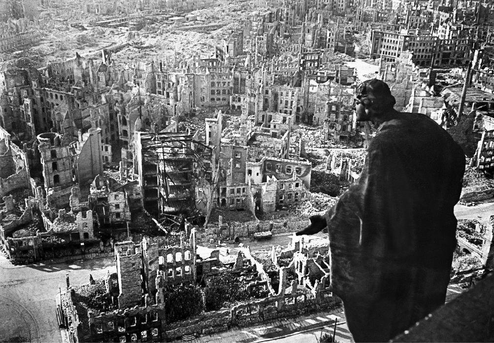 Fotografia em preto e branco. Vista aérea para uma cidade com edifícios destruídos e escombros. À direita, uma estátua de um santo em perfil, com os braços abertos.