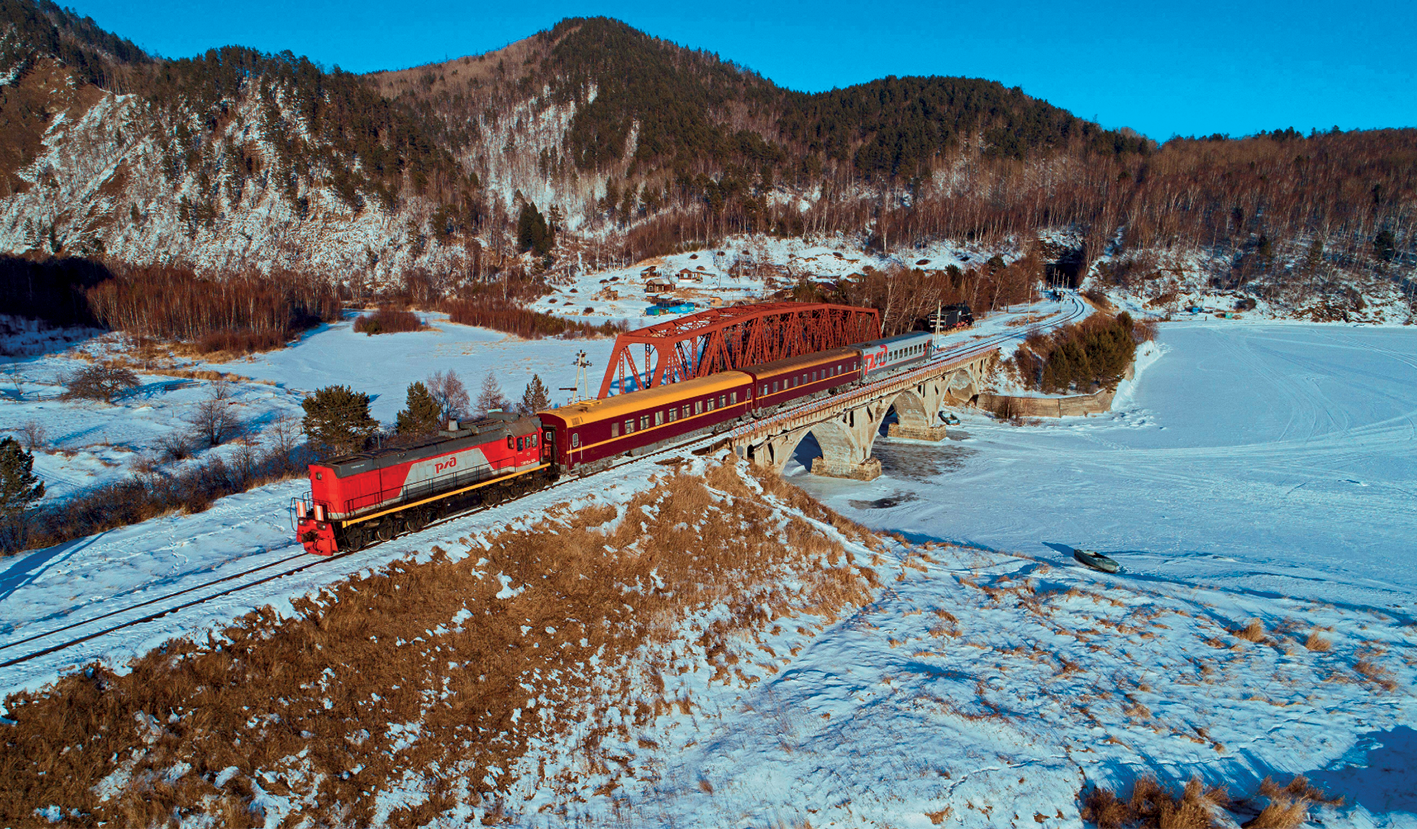 Fotografia. Vista de uma área recoberta de neve onde há uma ferrovia sobre uma ponte e um trem circulando sobre os trilhos. À direita, o rio sob a ponte está congelado e coberto de neve. As partes de terra possuem neve e alguma vegetação rasteiras. À esquerda, montanhas cobertas de neve e fragmentos florestais em suas vertentes com folhagem seca.