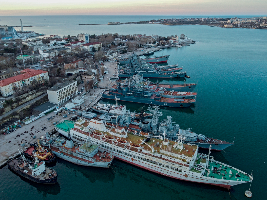 Fotografia. Destaque para a vista para uma base naval. À direita, o mar e embarcações de diversos tamanhos e tipos (militar, cruzeiro, pesqueiro) atracadas no cais. Ao fundo, porção do continente avançando no mar. À esquerda, via de circulação interna da base naval e edificações. Atrás da base, área urbana à beira-mar, cujo adensamento é esparso, arborizado e predominantemente horizontal.