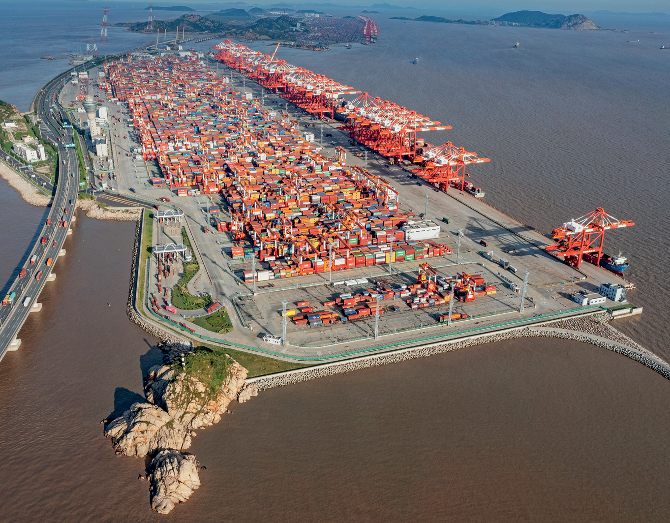 Fotografia. Vista aérea de uma zona portuária. No primeiro plano, vista de um porto construído sobre uma ilha, com uma concentração de contêineres empilhados em um extenso pátio. À esquerda, uma ponte usada para conectar a ilha ao continente. No segundo plano, ao fundo, vista de outras ilhas.