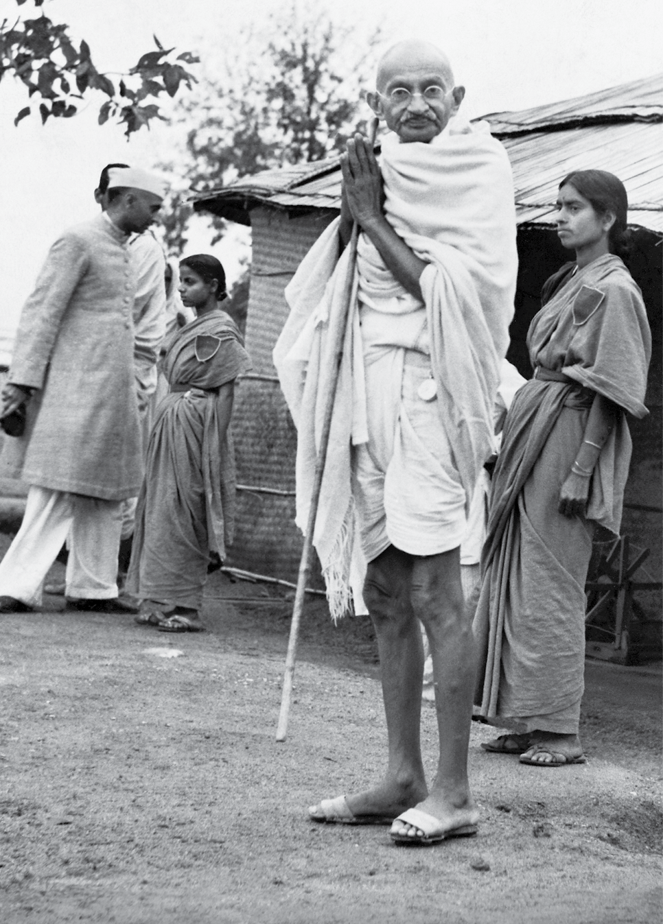 Fotografia em preto e branco. Em primeiro plano, destaque para Mahatma Gandhi. Ele está em pé, segura entre as mãos espalmadas um cajado de madeira, veste roupas brancas, óculos e olha em direção à câmera que registrou a imagem. Atrás dele, ao fundo, pessoas transitam. O chão é de terra batida.