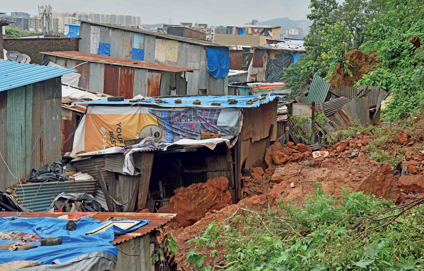 Fotografia. Vista de uma paisagem urbana. Na primeiro plano, diversas habitações precárias, construídas com materiais alternativos, como telhas de amianto, lonas e ripas de madeira, em um terreno íngreme e úmido. No segundo plano, ao fundo, observa-se alguns edifícios.