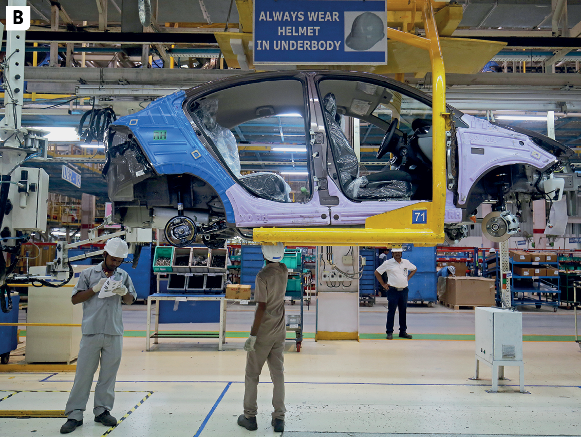 Fotografia B. Vista do interior de uma indústria automobilística. No primeiro plano, linha de montagem com a estrutura de um carro suspenso. Abaixo, dois operários uniformizados e de capacetes. No segundo plano, vista de outros setores da indústria.