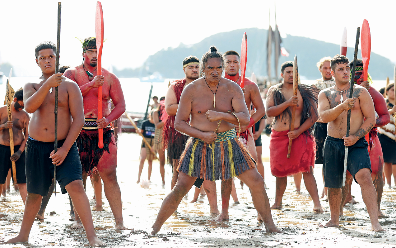 Fotografia. Na areia de uma praia, um grupo de homens está em pé com as pernas abertas, lado a lado, em três fileiras, uma atrás da outra. Alguns vestem bermuda, dois vestem uma saia e todos estão sem camisa e descalços. Alguns têm pinturas no corpo e seguram varas ou remos de barco na frente do corpo.