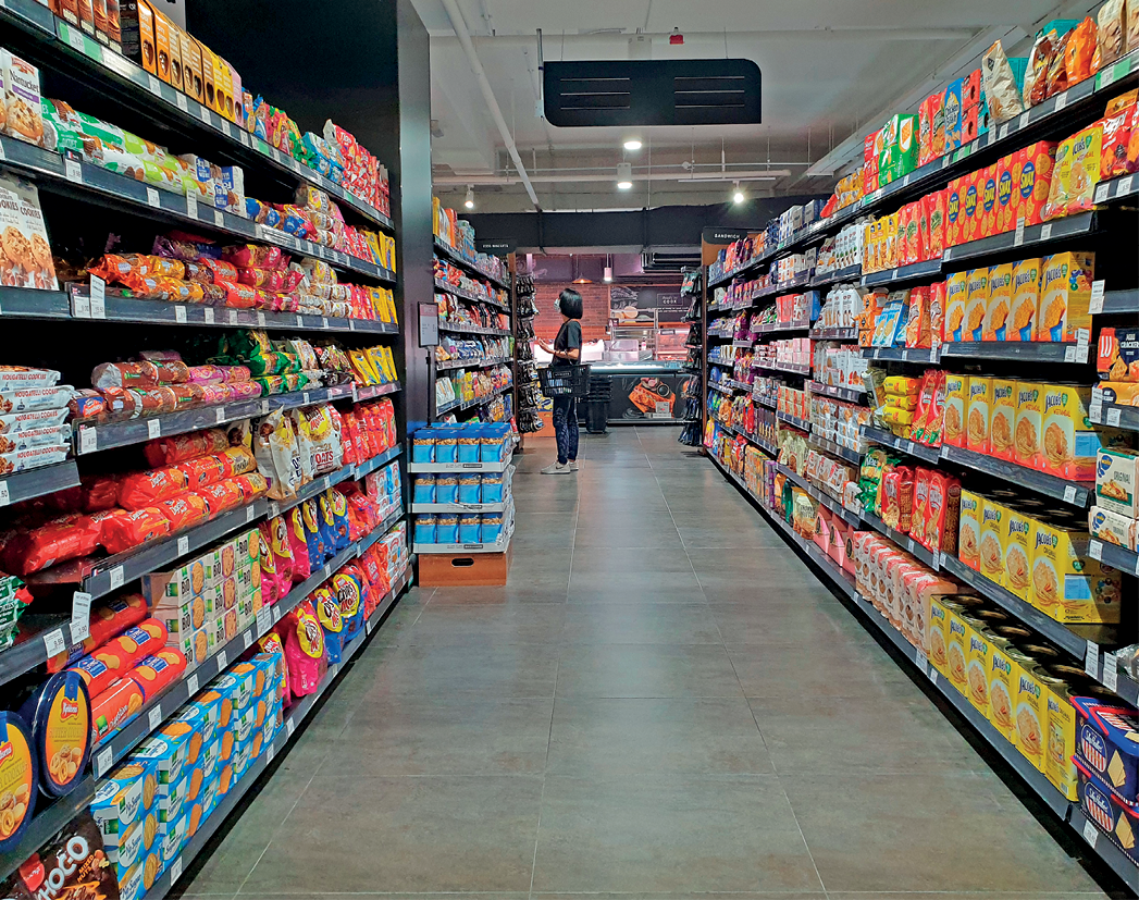 Fotografia. Vista de um corredor de supermercado com prateleiras abastecidas de produtos diversos. Ao fundo, uma pessoa de pé observa os produtos disponíveis.