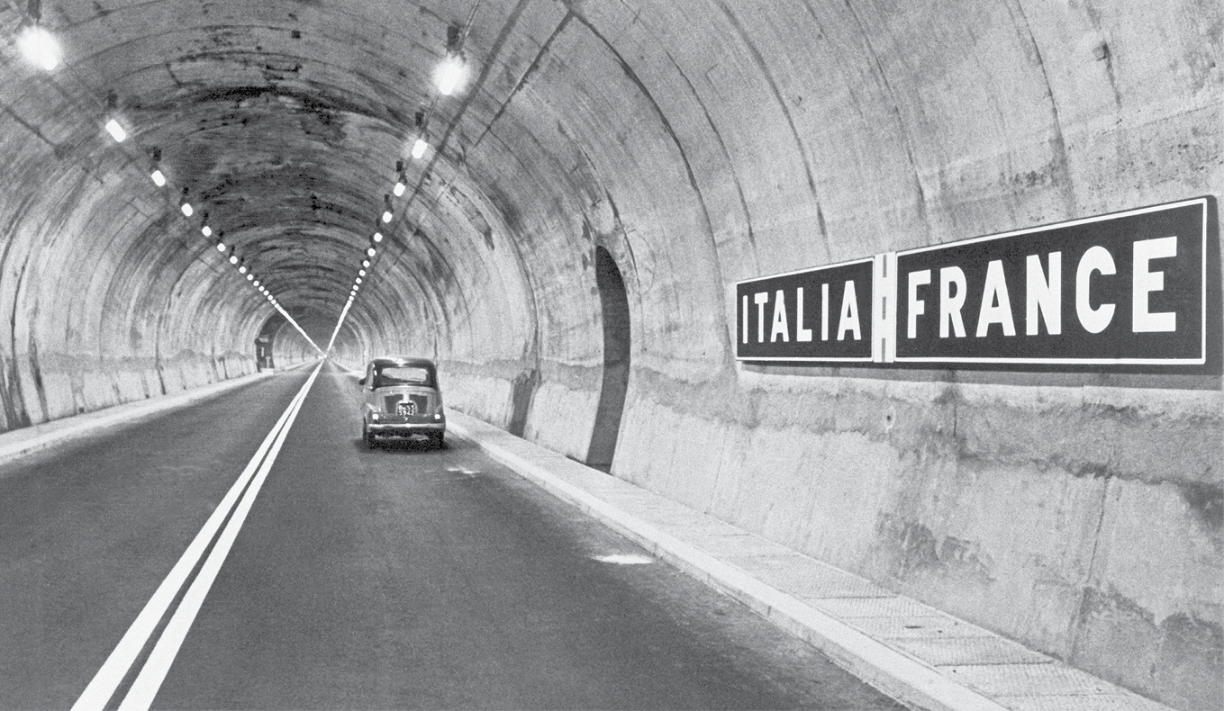 Fotografia em preto e branco. Destaque para uma pista de asfalto dentro um túnel de concreto. A pista é dupla e há um automóvel antigo circulando sobre a faixa da direita. Na parede direita do túnel, há duas placas: Italia e France. E no teto, há lustres com luzes acesas.