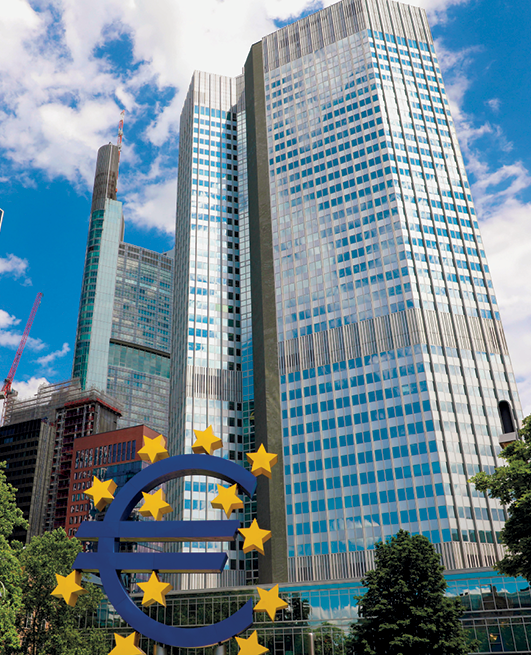 Fotografia. Vista para a fachada de conjunto de edifícios modernos e espelhados. À frente do prédio, o símbolo do euro, composto pela letra E, em azul, e 12 estrelas amarelas ao redor.