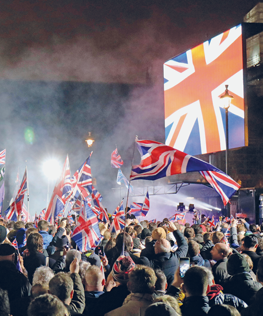 Fotografia. Vista para uma multidão de pessoas aglomeradas e de costas, segurando bandeiras do Reino Unido. Há fumaça sobre as pessoas. À direita, um telão com a imagem da bandeira do Reino Unido.