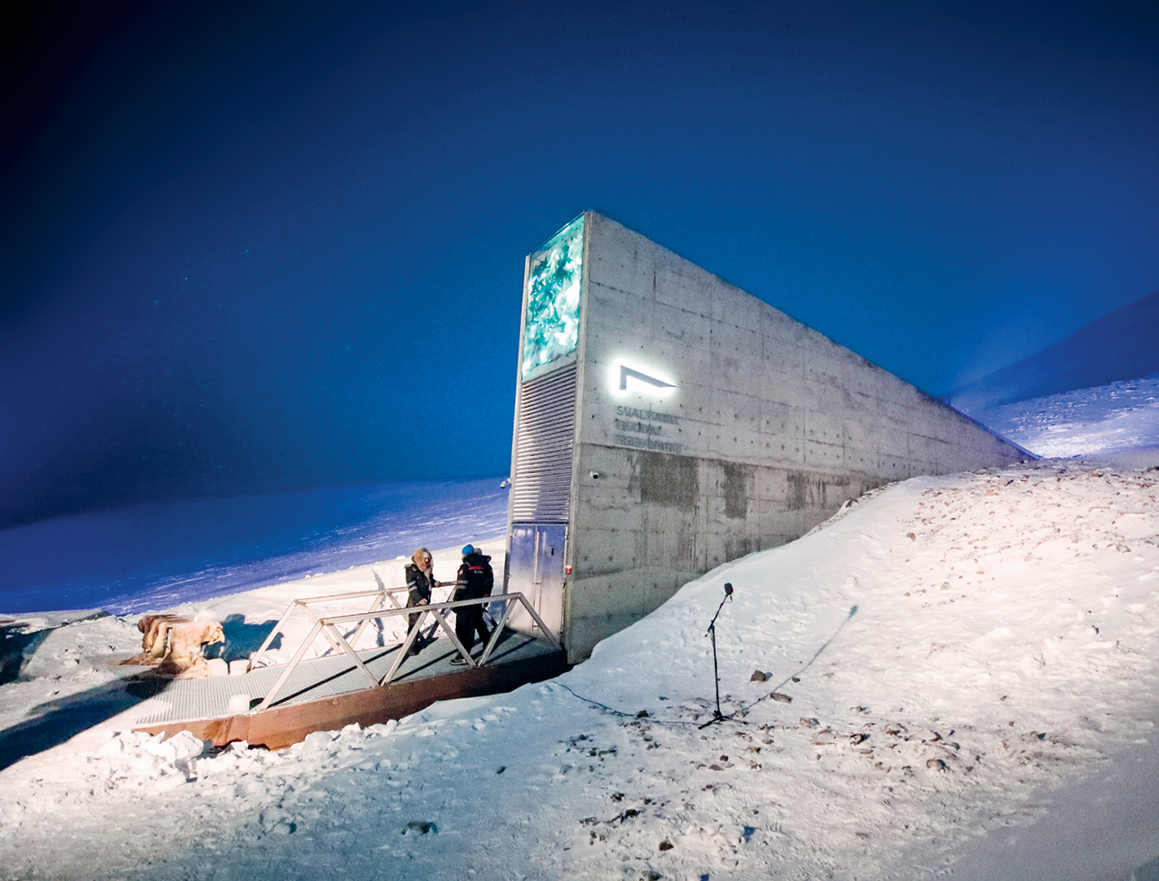 Fotografia. Vista para uma construção de concreto triangular com uma porta de vidro e uma passarela na entrada com duas pessoas em pé. Ao redor da construção, superfície inclinada coberta de neve e céu azul noturno.