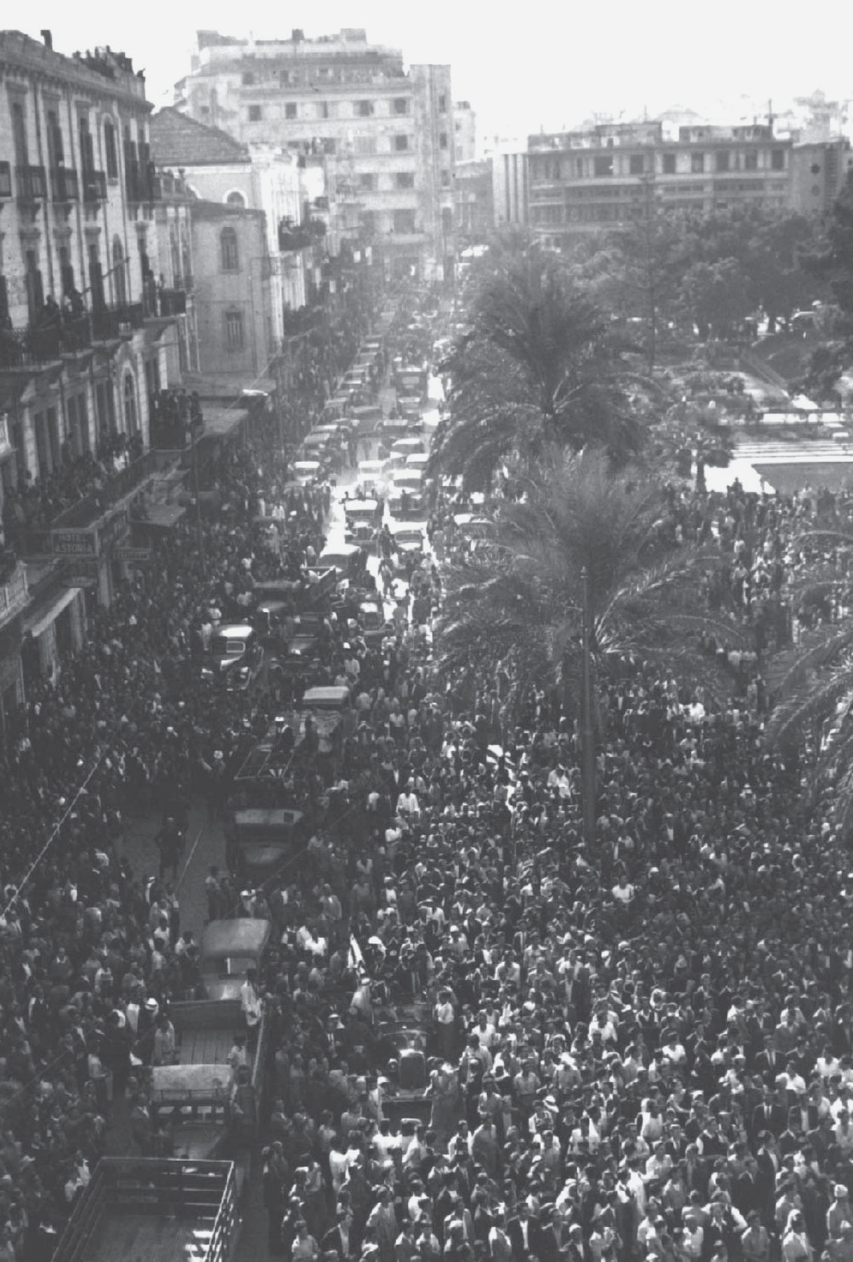 Fotografia em preto e branco. Vista de uma rua ocupada por uma multidão de pessoas e veículos. À esquerda, fachadas de edifícios. À direita, praça com palmeiras ocupada por pessoas. Ao fundo, rua com veículos, árvores e edificações.