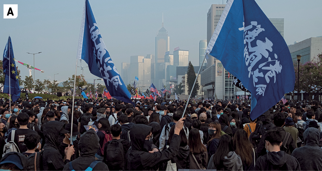Fotografia A. Vista para uma multidão de pessoas em um espaço público aberto. Algumas pessoas erguem bandeiras azuis. No fundo, edifícios e arranha-céus.