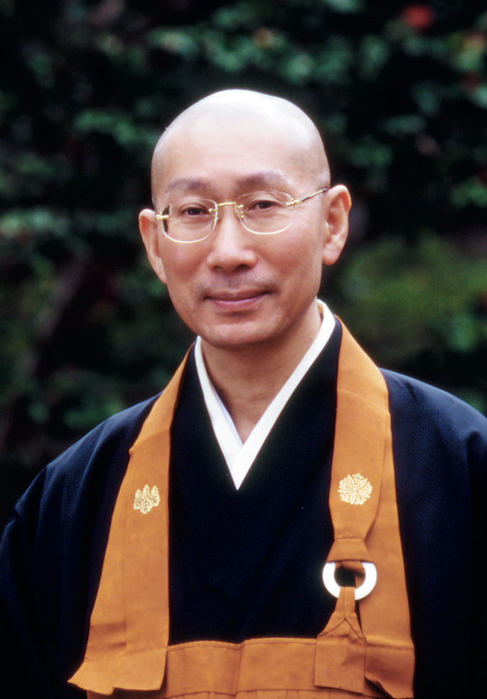 Fotografia. Retrato de um homem calvo e de óculos, vestindo quimono azul-escuro e avental marrom. Ao fundo, folhagem verde-escura.
