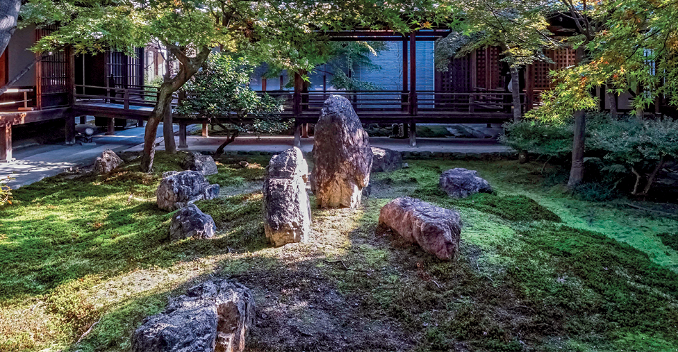 Fotografia. No centro, um jardim composto por gramado verde, alguns fragmentos de rocha no centro e árvores ao redor. Ao fundo, uma construção com pilares e uma pequena passarela ligando o jardim.
