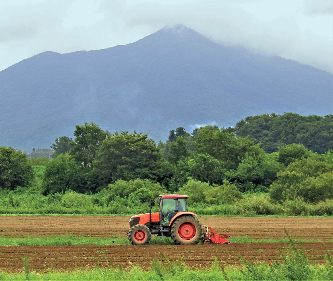 Fotografia. Imagem de um trator vermelho em primeiro plano com um implemento acoplado na parte de traz, em um terreno aplainado, com solo exposto e revolvido pelo maquinário. Em segundo plano, uma vegetação abundante. Em terceiro plano, uma montanha alto com nuvens no topo.