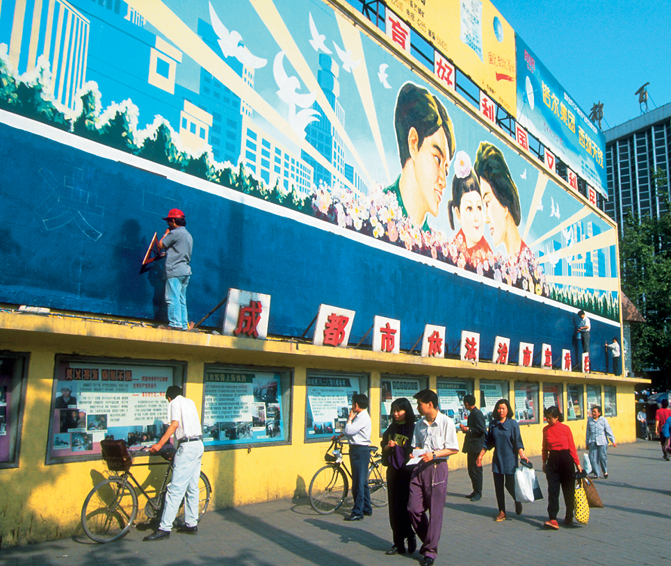 Fotografia. Pessoas caminham em uma calçada em um dia ensolarado. Acima, há um muro colorido, onde um homem está pintando alguns dizeres em mandarim. Mais acima, o desenho de uma família, composta por um homem, uma mulher e uma criança.
