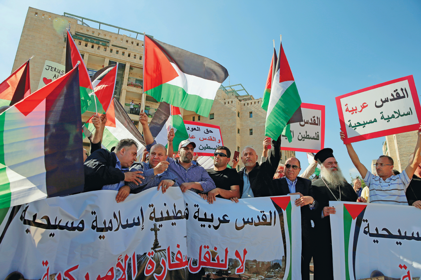 Fotografia. No primeiro plano, pessoas segurando uma grande faixa com escritos em árabe e, atrás delas, outras pessoas com bandeiras da Palestina e cartazes com escritos em árabe. Atrás, edifícios.