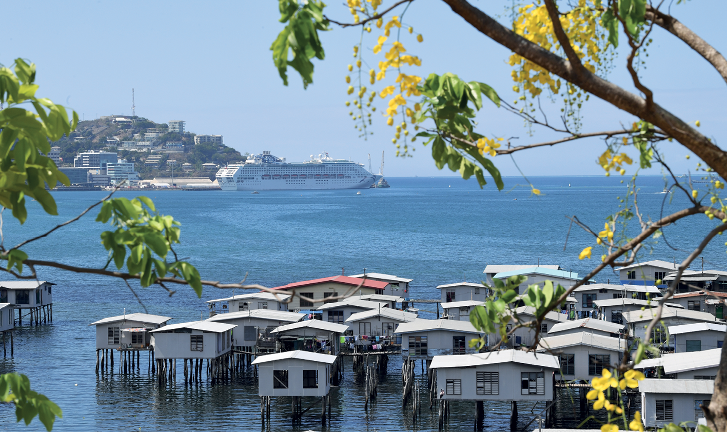 Fotografia. Em primeiro plano, agrupamento de casas suspensas sobre a água por meio de estacas. Ao fundo, o mar, um grande navio e um morro com construções em sua encosta. A paisagem é ensolarada e o céu está azul, sem nuvens.