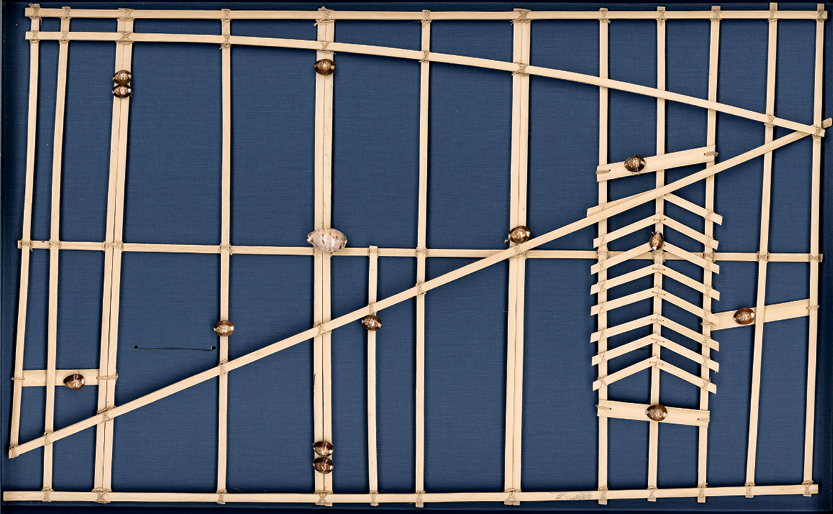 Fotografia. Mapa confeccionado com varetas de madeira que formam diversos retângulos e outras figuras geométricas. Em alguns pontos das varetas há conchas amarronzadas.