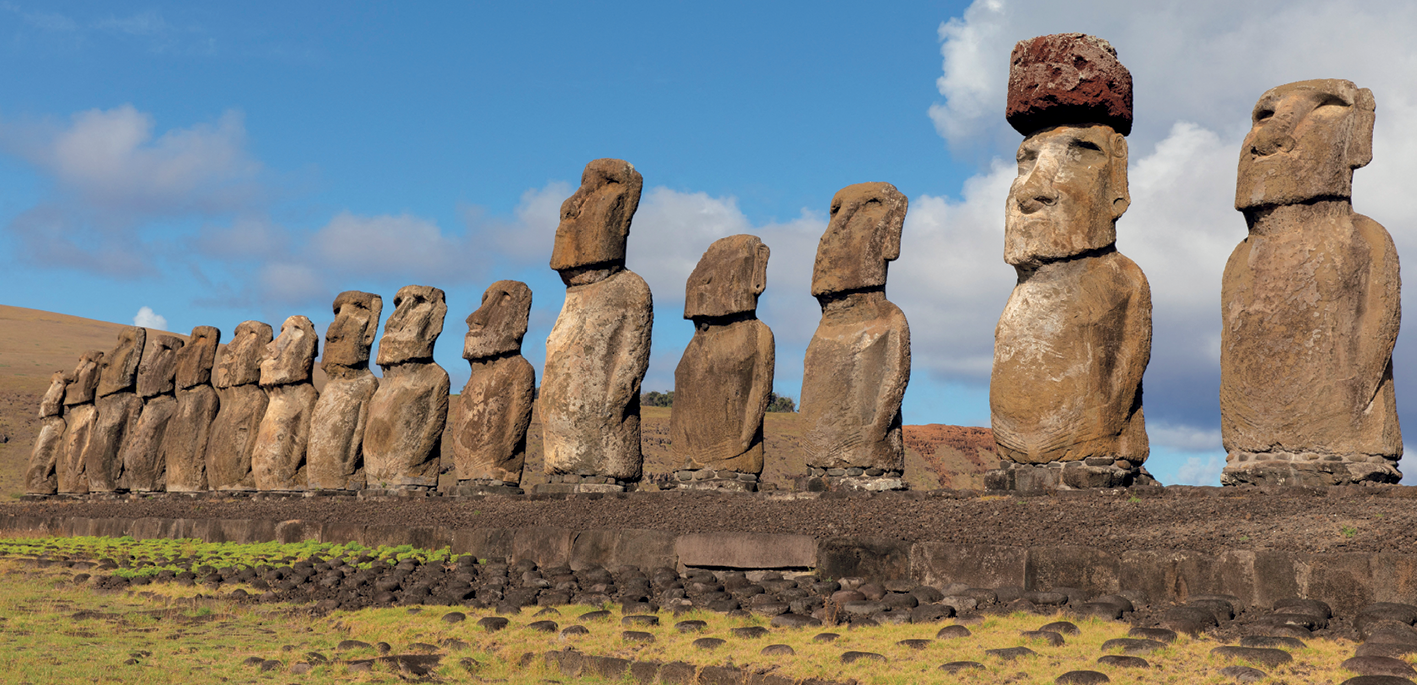 Fotografia. Imagem de um local aberto com diversas esculturas gigantes esculpidas em pedra. Elas estão enfileiradas lado a lado e têm formato humano. Ao fundo, céu azul.