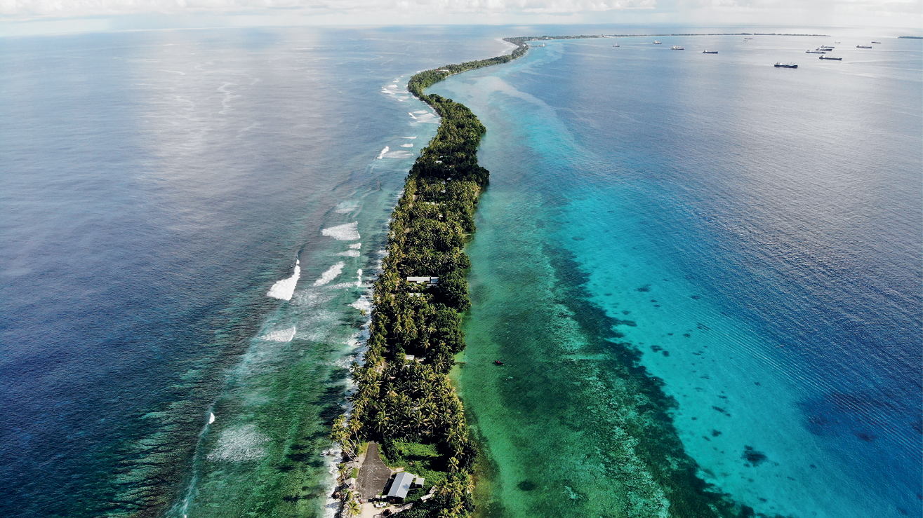 Fotografia. Uma estreita faixa de terra com poucas construções e bastante vegetação. Ao redor, o mar com tons de azul e verde. Ao fundo, alguns navios no mar.