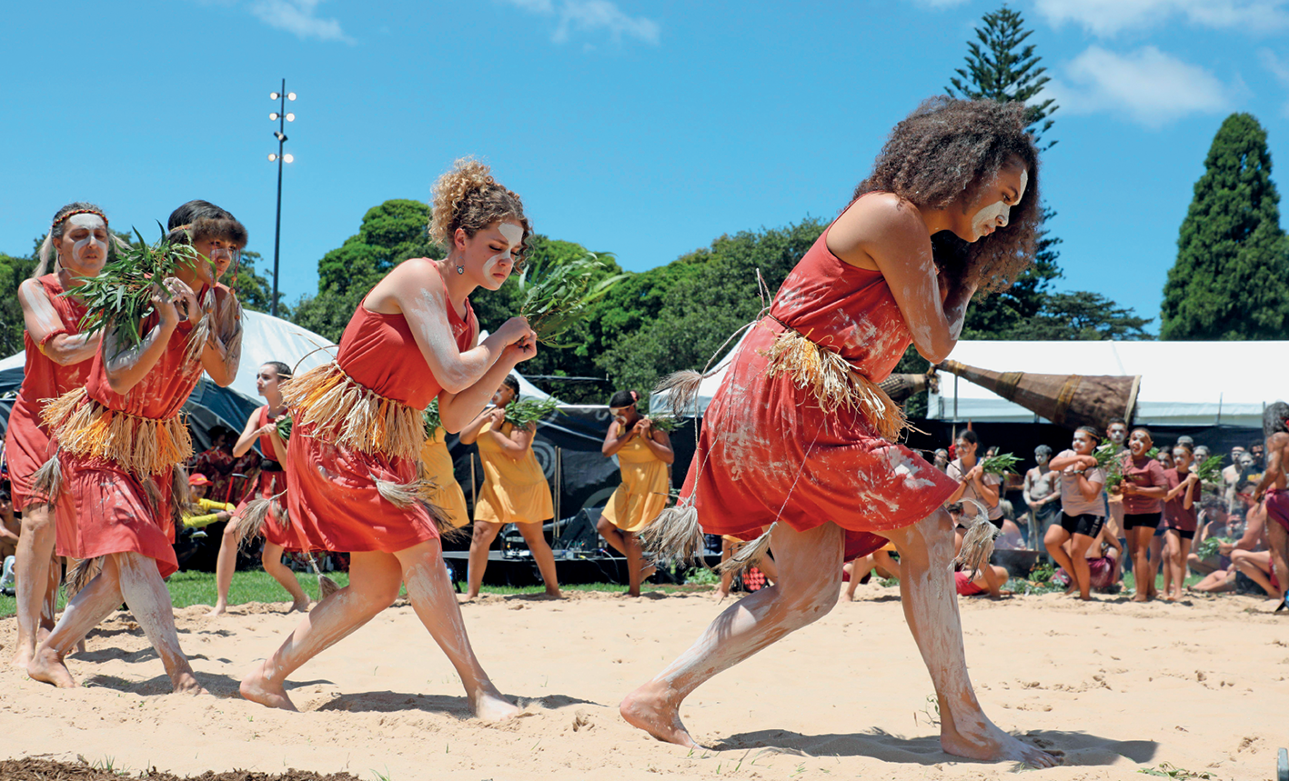 Fotografia. Grupo de mulheres enfileiradas uma atrás da outra, em semicírculo, dançando sobre chão de areia. Elas estão com o rosto pintado, descalças, vestem vestidos iguais, em tom de vermelho e com faixa de tufos de palha na cintura, na altura dos joelhos. Ao fundo, toldos, algumas pessoas observando e árvores elevadas de folhagem verde.