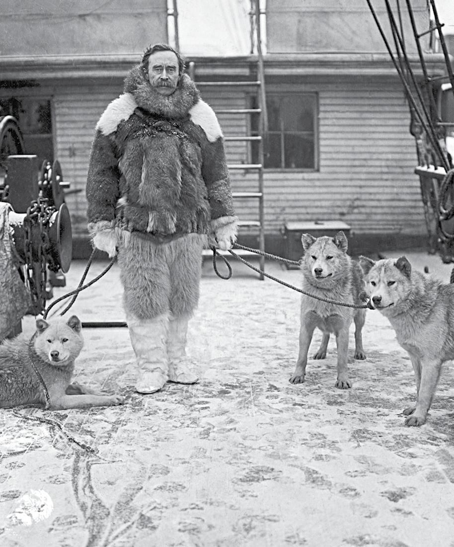 Fotografia em preto e branco. No primeiro plano, vista de um homem agasalhado com roupas de pele. O chão está coberto de neve. Junto a ele, três cachorros presos por uma guia. No segundo plano, vista de uma casa com uma escada.