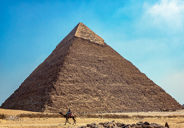 Fotografia. Vista frontal de uma construção em forma de pirâmide, na cor marrom escuro e topo mais claro.