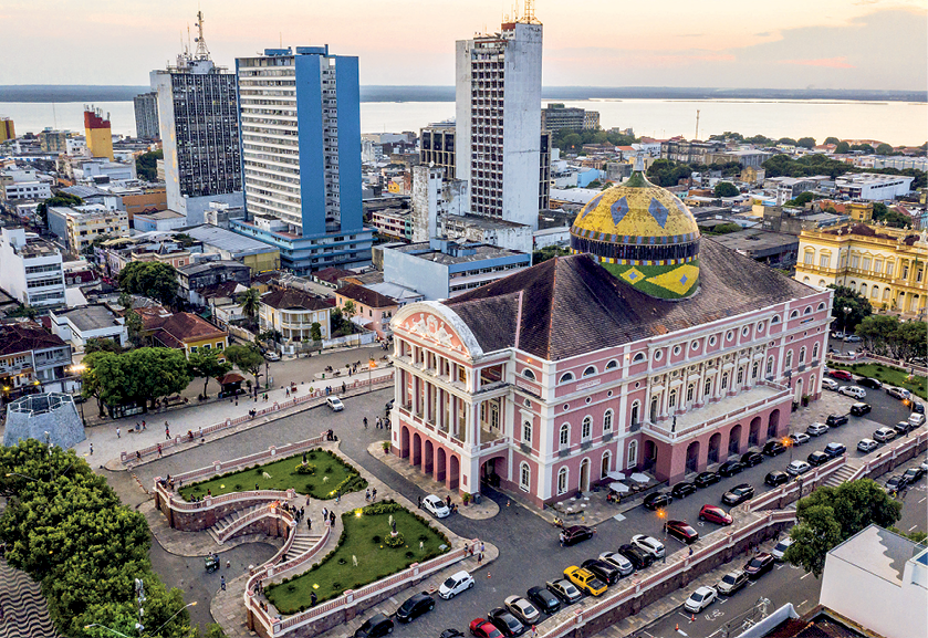 Fotografia. Ao centro, o Teatro Amazonas, uma grande construção que tem no topo uma área esférica. Na parte inferior esquerda, uma praça com gramado. Na parte inferior esquerda, veículos estacionados. Ao fundo, prédios, casas e o mar.