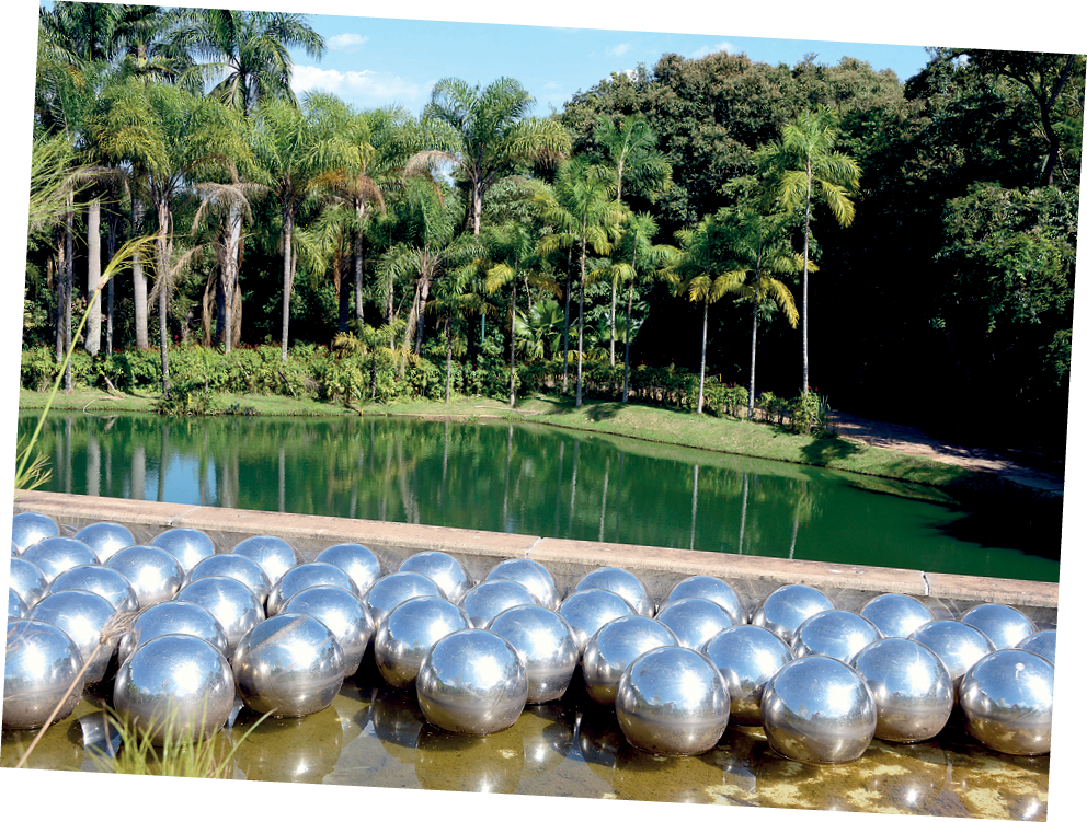 Fotografia. Diversas esferas prateadas sobre região espelhada. Ao fundo, um lago, vegetação e árvores.