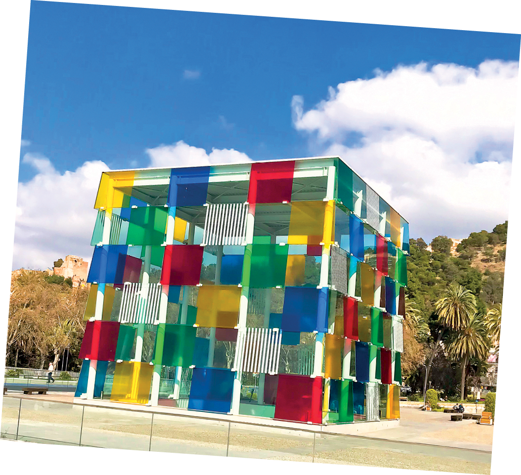 Fotografia. Construção de vidro em formato de cubo. Os quadrados dos cubos estão pintados nas cores amarela, vermelha e azul intercaladas.