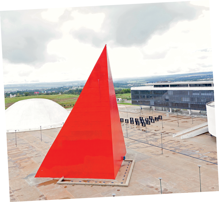 Fotografia. Vista do alto de construção vermelha em formato de pirâmide. À direita, construção baixa retangular com janelas de vidros. Ao fundo, grama e construções.