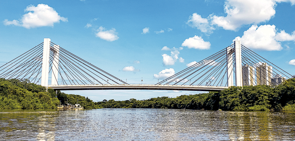 Fotografia.
A fotografia destaca a vista da lateral de uma ponte de ligação intermunicipal, composta por cabos que, com a base da ponte, lembram triângulos.
Abaixo da ponte destaca-se um rio e vegetação.
