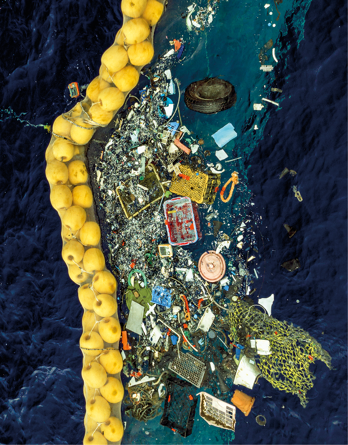 Fotografia. Vista aérea de uma área do mar e ao centro lixo plástico. A esquerda do lixo está um dispositivo desenvolvido para coleta de lixo plástico.