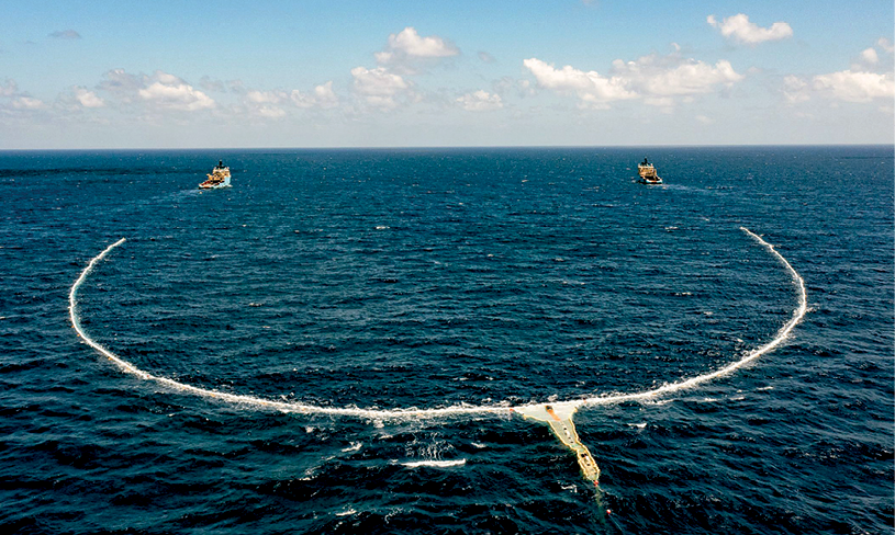 Fotografia. Vista aérea de uma região do mar em que aparece um dispositivo de coleta de lixo plástico sendo puxado nas suas duas extremidades por barcos.