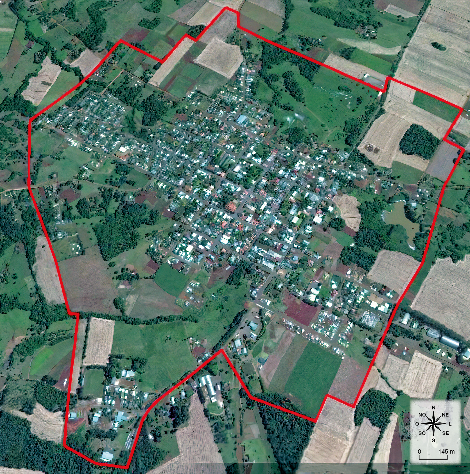 Fotografia.
Vista aérea de um município com construções e vegetação ao redor. Uma linha vermelha contorna o limite do município.
