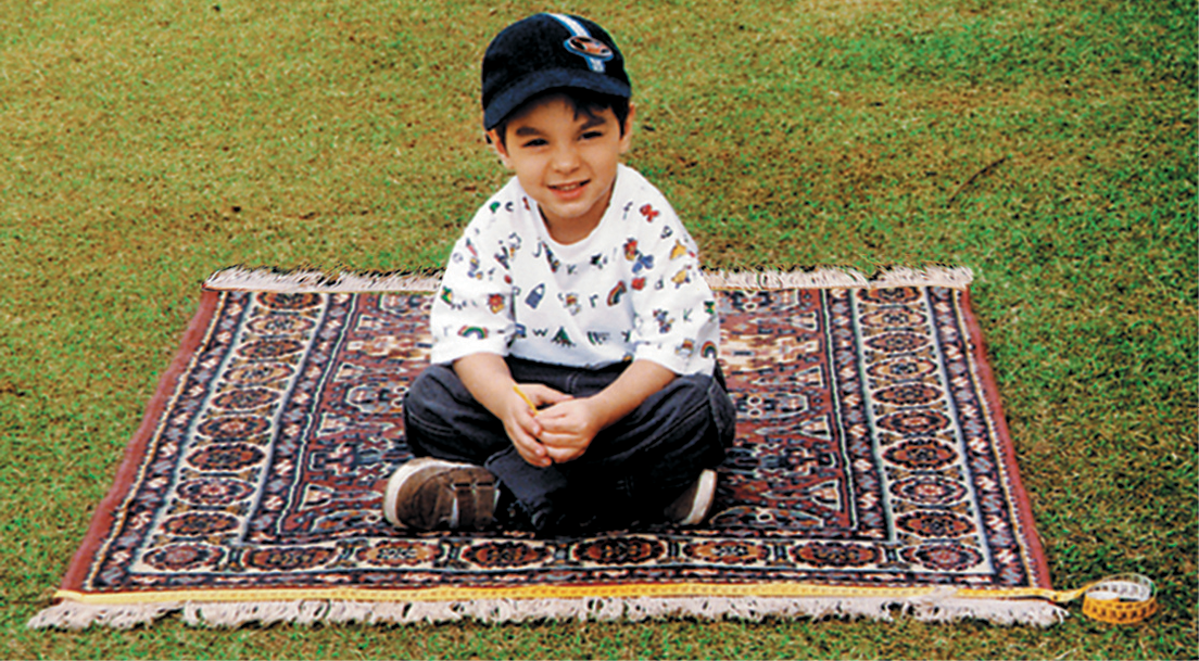 Fotografia.
Criança de boné, camisa branca estampada e calça preta. Está sentada em um tapete estampado, sob um gramado.
Há uma fita métrica indicando que o lado do tapete mede 1 metro.