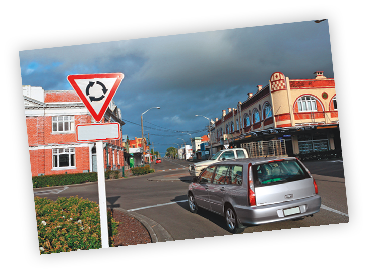 Fotografia. Carros trafegando em uma via pública. À esquerda, placa em formato triangular.