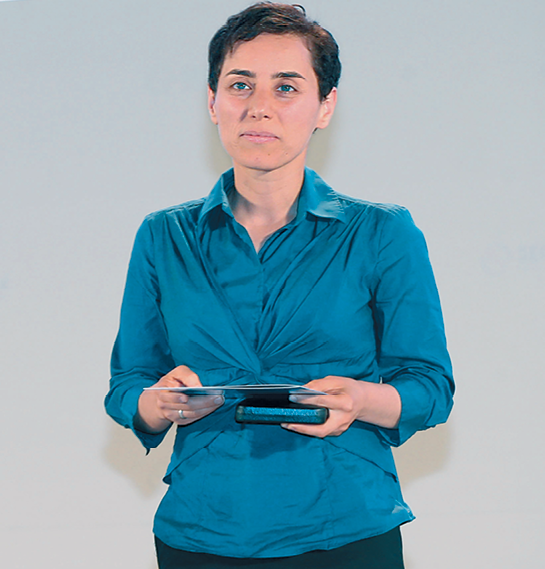 Fotografia. Mulher branca, de cabelos castanhos curtos, vestindo camisa azul. Ela segura alguns objetos nas mãos.