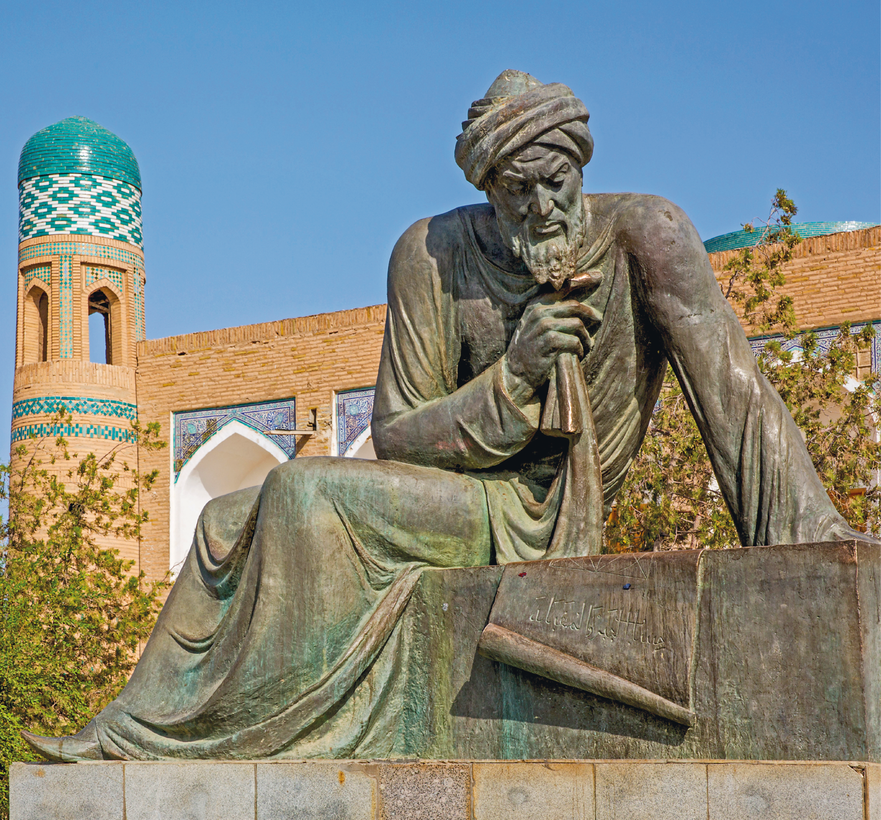 Fotografia. Estátua de um homem árabe de barba, turbante e túnica. Ele está sentado em um patamar com a cabeça para baixo olhando para um papiro aberto. Ao fundo, construção de tijolos com topo em detalhes verdes e brancos.