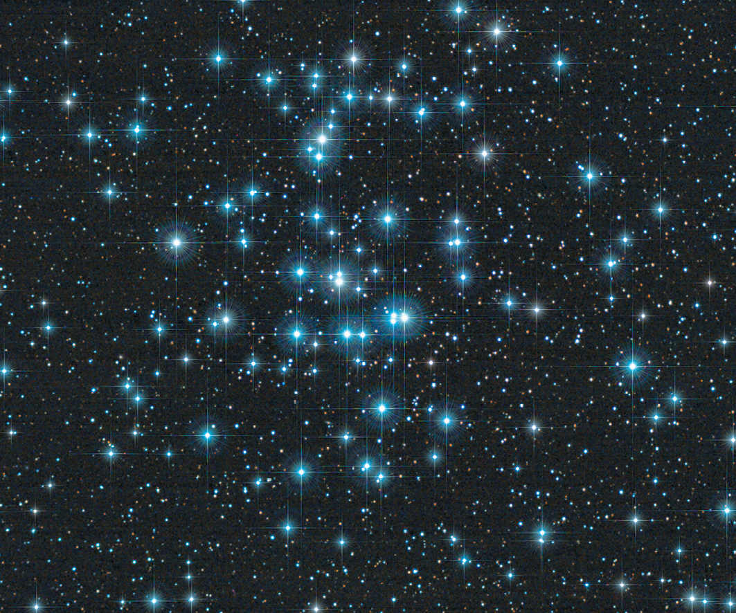 Fotografia.
Diversas estrelas de coloração azul em um céu noturno. No centro, se observa um agrupamento maior de estrelas.