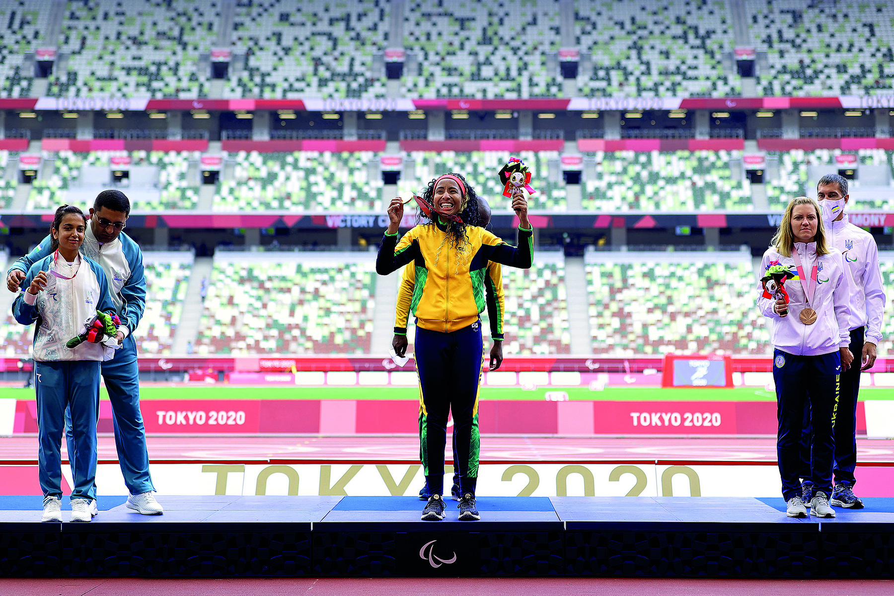 Fotografia. Pódio para recebimento de medalhas nos jogos paralímpicos de Tóquio. Ao centro encontra-se a atleta brasileira, ela segura na mão direita a medalha de ouro e na mão esquerda um buquê de flores. A sua direta está a atleta que ganhou a medalha de prata, ela também segura nas mãos a medalha e o buquê. A sua esquerda se encontra a medalhista de bronze, em sua mão direita o buquê. A medalha de bronze está pendurada no pescoço. Atrás delas se encontram seus respectivos assessores. Em segundo plano, a arquibancada com uma faixa vermelha e os dizeres: Tóquio 2020.