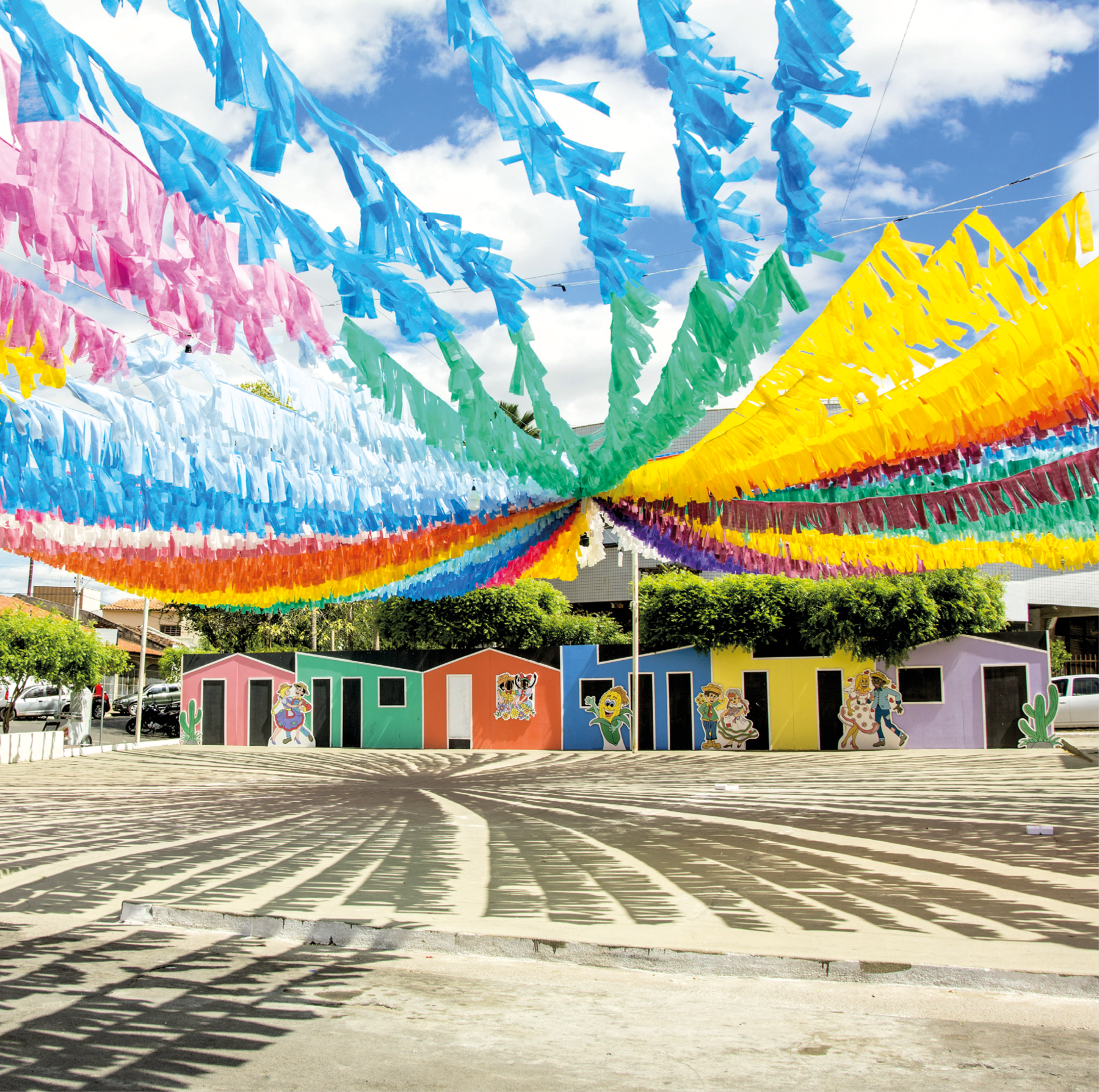 Fotografia. Vista parcial de praça com diversas fileiras de bandeirinhas coloridas penduradas. Ao fundo, casas coloridas e árvores