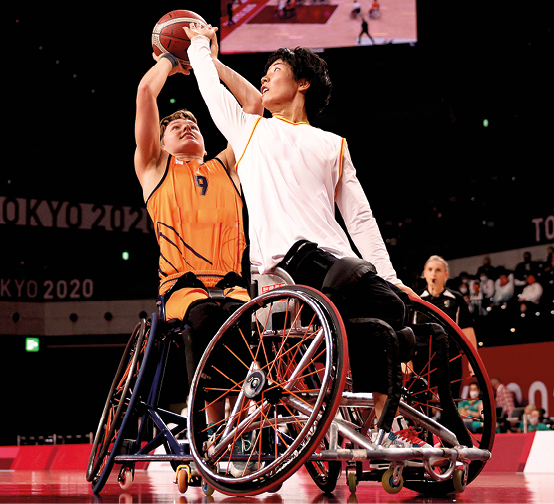 Fotografia. Destaque para dois homens em cadeira de rodas disputando uma bola de basquete. Ao fundo, arquibancada com pessoas.