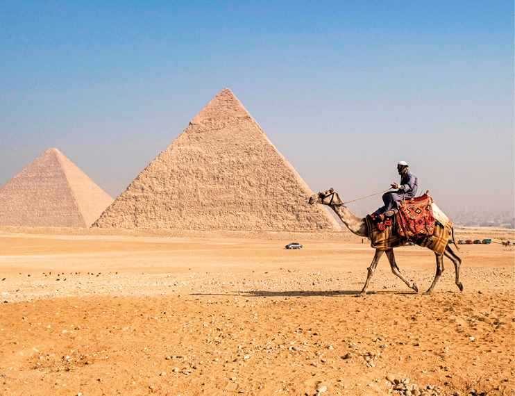 Fotografia. 
Vista frontal de pirâmides em solo de terra. À direita, homem sobre um camelo.