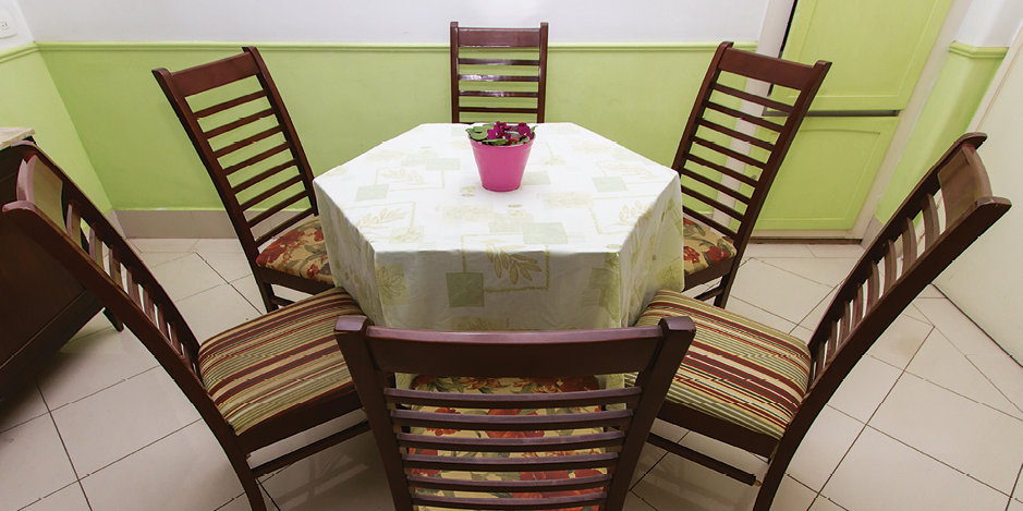 Fotografia. Vista do alto de mesa com tampo de formato hexagonal, com 6 cadeiras de madeira ao redor. Por cima da mesa está estendida uma toalha branca com decorações em tom claro e, ao centro, há um vaso rosa com folhas dentro.