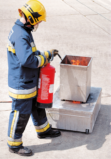 Fotografia. Pessoa de capacete amarelo e uniforme azul com faixas amarelas. Ela segura um extintor de incêndio vermelho, apontando para um recipiente de metal com fogo dentro.